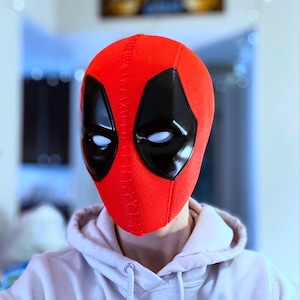 May include: A red Deadpool mask with black eye coverings and white eye details. The mask is made of a textured fabric and features visible stitching along the top and back. The mask is worn over a light-colored hoodie.