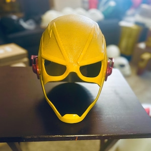 May include: A yellow superhero helmet with a textured surface and black eye openings. The helmet has red ear pieces and is sitting on a dark brown table. The helmet is a cosplay item.