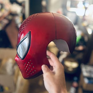 May include: A red, glossy mask with a smooth finish and a series of small, evenly spaced holes. The mask has a reflective, black eye covering and is held by a hand. The mask is likely a costume or cosplay item.