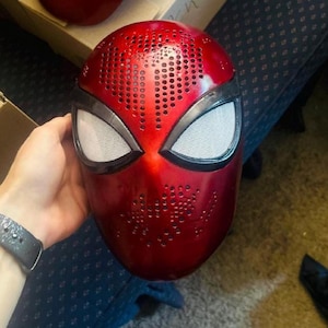 May include: A red Spider-Man mask with white eye lenses and a perforated design. The mask is a vibrant red with black accents around the eyes. The image shows the mask held by a person.