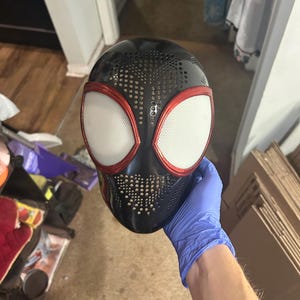 May include: Black and red Spider-Man mask with white eye lenses. The mask has a perforated design on the top and sides. The eye frames are red, and the lenses are white. The mask is held by a person wearing a blue glove.