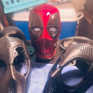 May include: A red and black Deadpool mask is the focal point, surrounded by several black masks. The red mask has a glossy finish with white eye openings and a mesh mouth area. The other masks are matte black, with various designs and textures.
