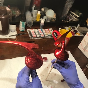 May include: Two glossy red musical note ornaments are held. The ornaments are on wooden sticks, and appear freshly painted. The background shows art supplies and a red mug. The ornaments are likely for decoration.