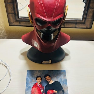 Pode incluir: Uma máscara vermelha e amarela do Flash com um design de raio. A máscara está assinada "Grant Gustin". Uma foto de duas pessoas, uma delas vestindo um traje vermelho e amarelo do Flash, segurando uma máscara vermelha e amarela do Flash.