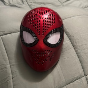 May include: A red Spider-Man mask-shaped speaker with black detailing around the eyes. The mask features many small holes for sound and white mesh over the eye openings. The speaker is on a grey, quilted surface.