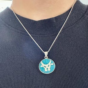 May include: Silver pendant necklace featuring a round turquoise stone with a white bull design. The pendant hangs from a delicate silver chain. The necklace is worn against a dark blue shirt.