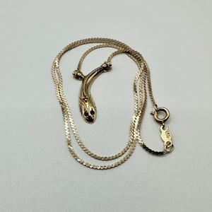 May include: A gold-toned snake pendant necklace. The snake head features two red gemstones for eyes. The chain is a delicate, flat design with a spring ring clasp. The necklace is displayed on a white surface.