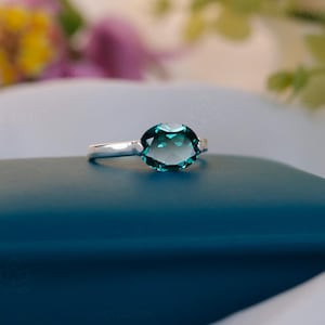 Puede incluir: Un anillo de plata con una gran gema ovalada de color verde azulado. El anillo está sobre una superficie azul oscuro, con flores borrosas en el fondo. La gema tiene un aspecto facetado, captando la luz. El anillo es una joya.