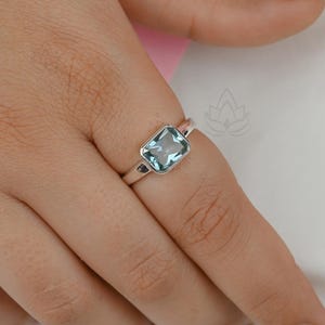 Op de afbeelding: Een zilveren ring met een rechthoekige, lichtblauwe edelsteen. De ring bevindt zich aan een vinger en toont het ontwerp en de kleur van het sieraad. De edelsteen is gezet in een eenvoudige, elegante band.