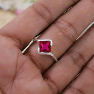 May include: A silver ring with a square shaped pink gemstone. The ring is on a person's finger.