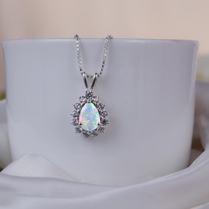 May include: A silver pendant necklace featuring a pear-shaped opal gemstone surrounded by a halo of small, clear gemstones. The necklace is displayed on a white cup, set against a soft, white fabric background.