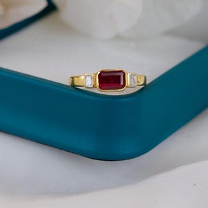 May include: A gold ring with a rectangular, deep red gemstone center and two smaller white gemstones on either side. The ring is displayed in a teal jewelry box, with white fabric and flowers in the background.