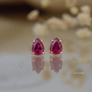 Puede incluir: Un par de pendientes de oro rosa con un rubí en forma de pera. Los pendientes se colocan sobre un fondo suave y borroso, resaltando el color intenso y el brillo de los rubíes.