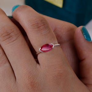 May include: A silver ring featuring a marquise-cut, vibrant red gemstone. The ring's band is delicate, and the gemstone is set in a minimalist style. The ring is displayed on a hand with turquoise nail polish.