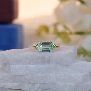 Puede incluir: Un anillo de oro con una piedra preciosa verde claro con talla esmeralda. El anillo está colocado sobre una superficie de mármol blanco, con un fondo borroso de flores blancas y otros objetos. El anillo tiene un diseño sencillo y elegante.