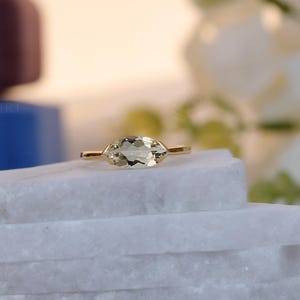 Può includere: Un anello d'oro con una gemma ovale verde chiaro. L'anello è appoggiato su una superficie di marmo bianco. Lo sfondo include elementi sfocati di blu, viola e bianco.