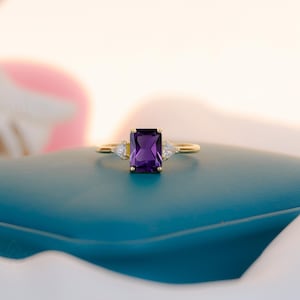 May include: A gold ring featuring a rectangular, deep purple gemstone, with two smaller, triangular white gemstones either side. The ring is displayed on a teal surface, with a soft, blurred background.