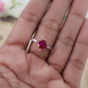 Peut inclure: Une bague en argent ornée d'une pierre précieuse rouge vif de forme carrée. La bague est montée sur un fin anneau, mettant en valeur la couleur et la forme de la pierre. La bague est présentée sur une main.