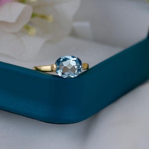Könnte beinhalten: Ein goldener Ring mit einem großen, ovalen, hellblauen Edelstein. Der Ring ist in einem goldenen Band gefasst und wird in einer türkisfarbenen Schmuckschatulle mit weißem Innenraum präsentiert. Blumen sind im Hintergrund zu sehen.