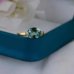 May include: A gold ring with a large, oval-cut, light teal gemstone. The ring is set in a gold band and rests on a teal blue jewelry box. The background includes white fabric and flowers.
