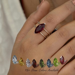 May include: A silver ring with a large, marquise-cut, deep red gemstone. The ring has a double-band design. The image also displays a variety of gemstone colors, with the text "15+ Stone Colors Available".