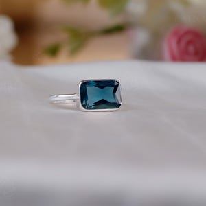Könnte beinhalten: Ein silberner Ring mit einem rechteckigen, dunkelblauen Edelstein. Der Ring liegt auf einer weißen Oberfläche, mit unscharfen Blumen im Hintergrund. Der Edelstein hat eine tiefe, gesättigte Farbe und der Ring ein schlichtes, elegantes Design.