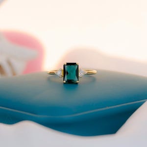 Puede incluir: Un anillo de oro con una piedra preciosa rectangular de color verde oscuro como foco principal. Dos piedras blancas triangulares más pequeñas flanquean la piedra principal. El anillo está sobre una superficie azul turquesa, con un fondo suave y borroso.