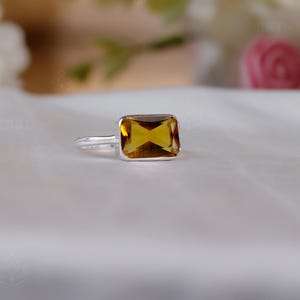 Op de afbeelding: Een zilveren ring met een rechthoekige, amberkleurige edelsteen. De edelsteen is gezet in een eenvoudig, elegant ontwerp. De ring wordt gepresenteerd op een wit oppervlak, met zachte bloemelementen op de achtergrond. De ring is een sieraad.
