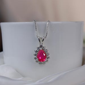 Puede incluir: Un collar con colgante de plata con una piedra preciosa rosa brillante en forma de lágrima, rodeada de piedras más pequeñas y transparentes. El colgante cuelga de una delicada cadena. El collar se exhibe sobre una superficie blanca.