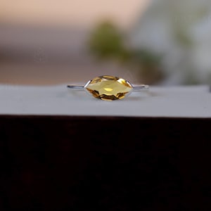 Peut inclure: Une bague en argent ornée d'une pierre jaune de forme marquise. La pierre est sertie sur un anneau simple et élégant. La bague est présentée sur une surface blanche à l'intérieur d'un coffret marron foncé, avec un arrière-plan flou.