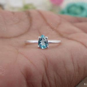 Op de afbeelding: Een zilveren ring met een peervormige, lichtblauwe edelsteen. De edelsteen is het middelpunt, gezet op een slanke, gepolijste zilveren band. De ring wordt getoond tegen een huidkleurige achtergrond, wat het ontwerp van de sieraden benadrukt.