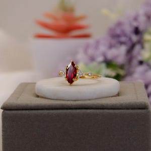 Peut inclure: Une bague en or ornée d'une pierre rouge foncé taillée en marquise. La bague est ornée de plus petites pierres claires de chaque côté de la pierre principale. Elle est présentée sur une pierre blanche, sur fond flou.
