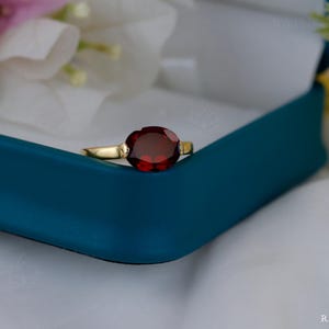 Peut inclure: Une bague en or avec une grosse pierre précieuse rouge foncé de coupe ovale. La bague est sertie dans un anneau en or et est présentée dans un écrin bleu sarcelle. Le fond comprend du tissu blanc et des accents floraux.