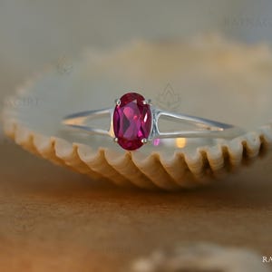 Peut inclure: Une bague en argent ornée d'un rubis ovale. La monture de la bague se divise en deux fines griffes qui maintiennent le rubis. La bague est présentée sur un fond neutre, mettant en valeur la couleur riche du rubis.