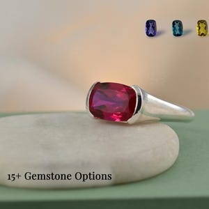 May include: A silver ring with a large, rectangular, deep red gemstone. Above the ring are five smaller gemstones in various colors: purple, teal, yellow, pink, and green. The text "15+ Gemstone Options" is visible. The ring is on a white stone.