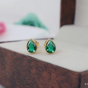 May include: A pair of stud earrings featuring teardrop-shaped, faceted green gemstones set in gold-toned metal frames. The earrings are positioned on a white surface within a brown textured box, displaying a clear, vibrant emerald-like hue.