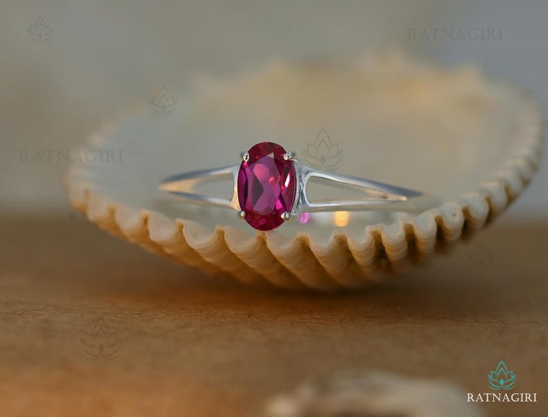 May include: A silver ring featuring a vibrant oval-cut ruby gemstone. The ring's band splits into two delicate prongs that cradle the ruby. The ring is set against a neutral background, highlighting the gemstone's rich color.