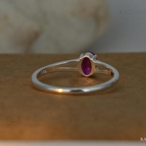 May include: A sterling silver ring with a vibrant oval-cut, deep pink gemstone. The ring features a split shank design, with the band separating near the gemstone. The gemstone is set in a bezel setting, and the ring is displayed on a brown surface.