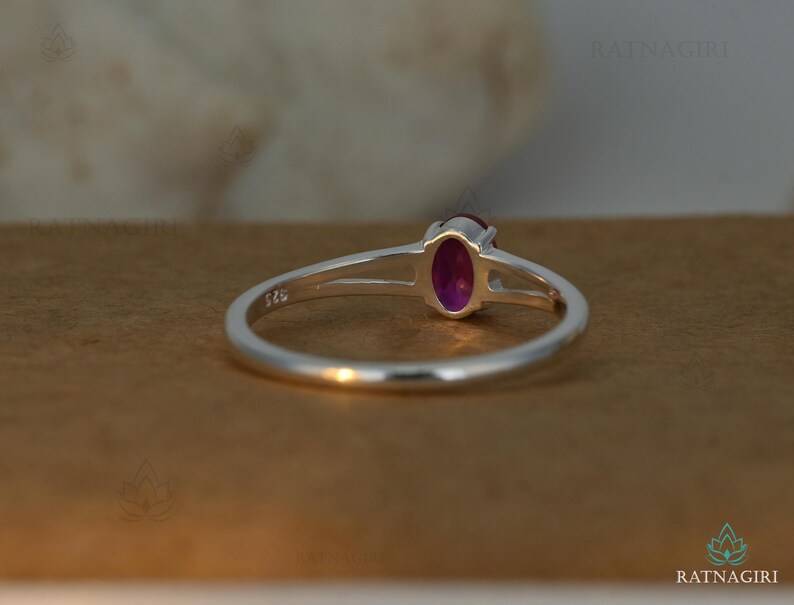 May include: A sterling silver ring with a vibrant oval-cut, deep pink gemstone. The ring features a split shank design, with the band separating near the gemstone. The gemstone is set in a bezel setting, and the ring is displayed on a brown surface.
