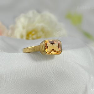 Puede incluir: Un anillo de oro con una piedra preciosa cuadrada de color naranja claro. El anillo tiene un diseño simple y elegante con una banda lisa y pulida. La piedra preciosa está engastada en un bisel. El anillo se muestra sobre tela blanca.