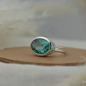Op de afbeelding: Een zilveren ring met een grote, ovaal geslepen, blauwgroene edelsteen in een gladde zetting. De ringband is gespleten in twee dunne, parallelle zilveren banen, met kleine, heldere, rond geslepen steentjes ingelegd aan de zijkanten.