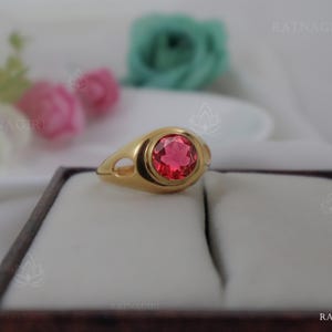 Könnte beinhalten: Ein goldener Ring mit einem großen, runden, roten Edelstein in einer Zargenfassung. Der Ring hat ein glattes, poliertes Band mit dekorativen Ausschnitten. Der Ring wird in einer braunen Schmuckschatulle präsentiert.
