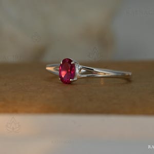 May include: A silver ring featuring a vibrant oval-cut ruby gemstone. The ring's band has a split design, adding a modern touch. The ruby is a deep, rich red color, set in a classic prong setting. The ring is displayed on a neutral background.