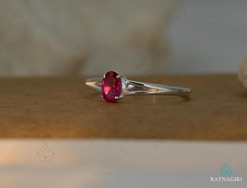 May include: A silver ring featuring a vibrant oval-cut ruby gemstone. The ring's band has a split design, adding a modern touch. The ruby is a deep, rich red color, set in a classic prong setting. The ring is displayed on a neutral background.