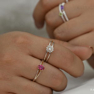 May include: Close-up of three rings on fingers. The rings are silver-toned. One ring has a cluster of small, clear stones. Another ring has a pink oval stone. A third ring has a purple stone and a green stone.