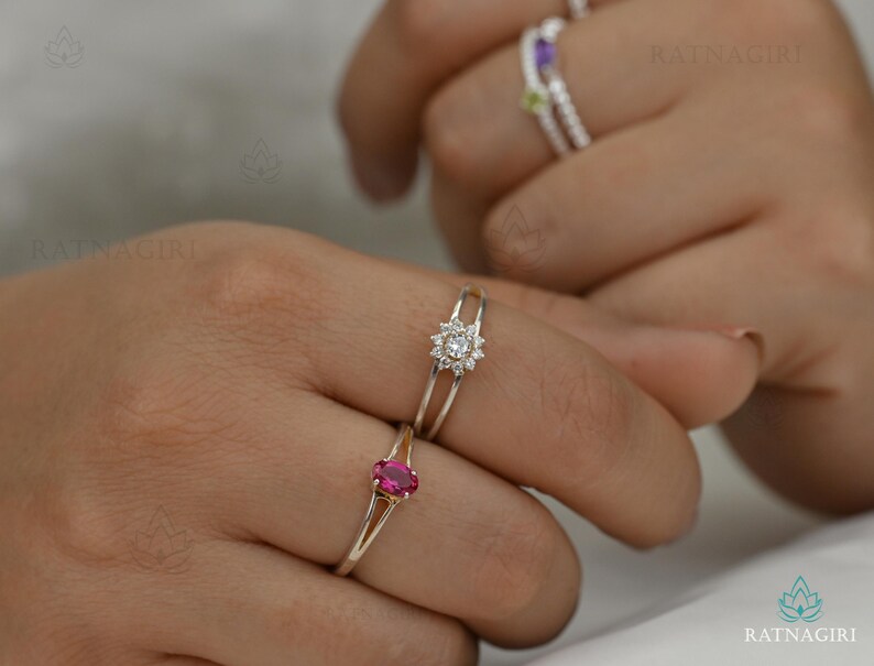 May include: Close-up of three rings on fingers. The rings are silver-toned. One ring has a cluster of small, clear stones. Another ring has a pink oval stone. A third ring has a purple stone and a green stone.