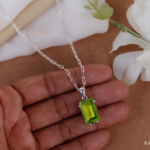 Puede incluir: Un collar de plata con un colgante rectangular de piedra preciosa verde claro. El colgante está engastado en un bisel de plata y cuelga de una delicada cadena de plata. El collar se muestra en una mano, con flores blancas en el fondo.