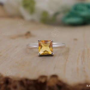 Può includere: Un anello d'argento con una gemma quadrata giallo dorato. L'anello è posto su una superficie di legno, con la parola "RATNAGIRI" visibile sullo sfondo. La gemma è il punto focale, mostrando il suo colore vibrante e il taglio.