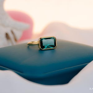 Op de afbeelding: Een gouden ring met een rechthoekige, smaragdgeslepen, donkergroene edelsteen. De ring ligt op een donkerblauwe ondergrond, met een zachte, vage achtergrond. De ring is een sieraad.