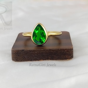 Puede incluir: Un anillo de oro con una piedra preciosa verde en forma de pera. El anillo está en un expositor de madera. RatnaGiri Jewels.